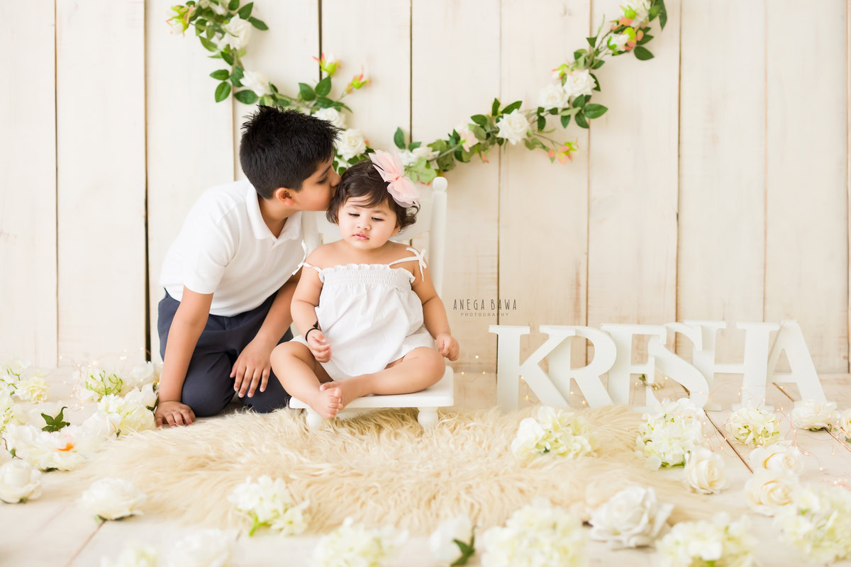 In a heartwarming moment captured during a baby pre-birthday photoshoot in Delhi, Anega Bawa, Gurgaon, and Noida, a 16-month-old girl shares a sweet kiss with her sibling. They sit together on a beige rug, surrounded by the warmth of their bond. Behind them, a leafy garland on the wall adds a touch of natural beauty to the scene. The beige backdrop provides a soft and neutral background, allowing the focus to remain on the adorable interaction between the siblings.