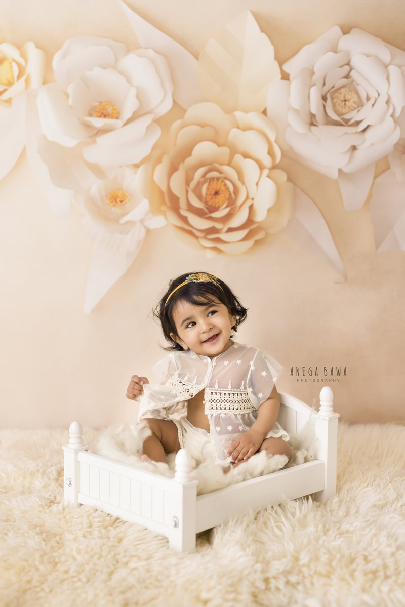 In Gurgaon and Delhi, Anega Bawa captures the charm of a 16-month-old girl seated on a white cot, surrounded by a beige rug against a backdrop adorned with delicate peach florals. Dressed in a cute net top and a lovely headband, she exudes innocence and sweetness, beautifully preserved in this photograph.