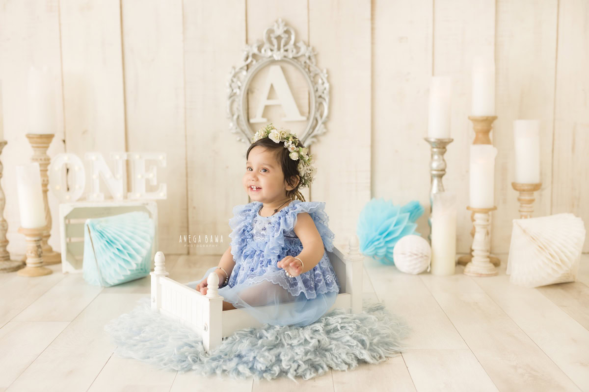 In this delightful toddler photoshoot captured by Anega Bawa, a 13-month-old girl sits comfortably on a white cot, surrounded by a serene ambiance. The blue rug beneath her adds a pop of color to the scene, complemented by playful blue pompoms. A candle stand adds a touch of warmth, while an alphabet frame adorning the wall provides a backdrop of education and learning. Against the neutral beige backdrop of Delhi, Gurgaon, or Noida, Anega Bawa expertly captures the innocence and charm of childhood in this enchanting photograph.
