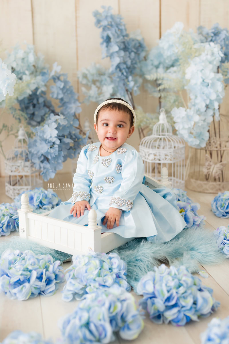 In this endearing toddler photoshoot by Anega Bawa, a 13-month-old girl sits gracefully on a white cot, surrounded by a whimsical atmosphere. Adorable castles and delicate blue flowers adorn the cot, adding a touch of fairy tale magic to the scene. The beige backdrop provides a soft and neutral background, allowing the focus to remain on the charming subject. Anega Bawa skillfully captures the innocence and beauty of childhood in this delightful photograph, showcasing the enchanting moments of toddlerhood in Delhi, Gurgaon, or Noida.