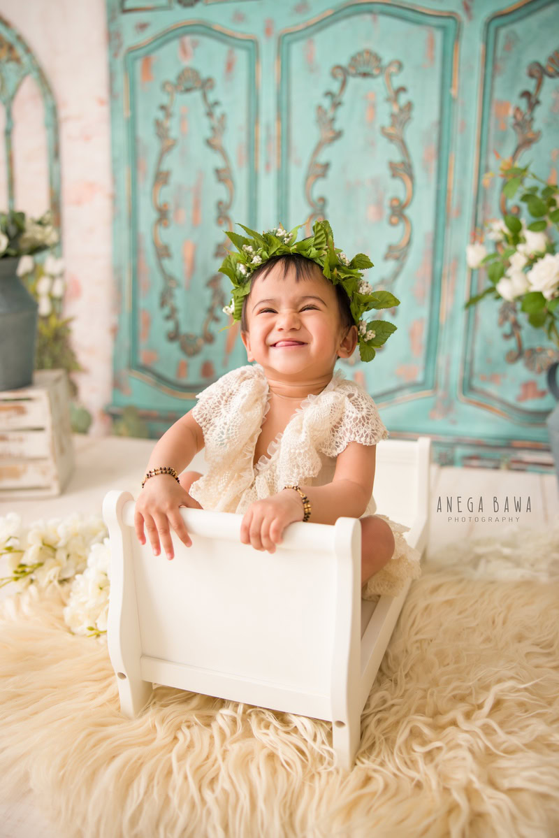 In this delightful photo session by Anega Bawa in Delhi, Gurgaon, a 16-month-old girl sits gracefully on a white cot, wearing a tiara band. Against a serene green backdrop, she exudes innocence and charm. The scene is completed with a cozy beige rug, adding a touch of comfort to the ambiance.