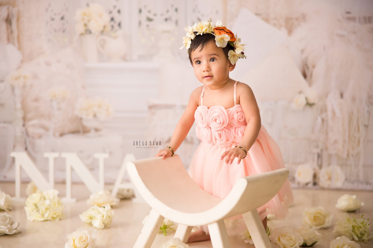 In Gurgaon and Delhi, Anega Bawa captures the grace of a 16-month-old girl standing beside a curvy chair, surrounded by ivory flowers. Adorned in a pink dress and a tiara band, she radiates elegance and charm, beautifully preserved in this photograph.
