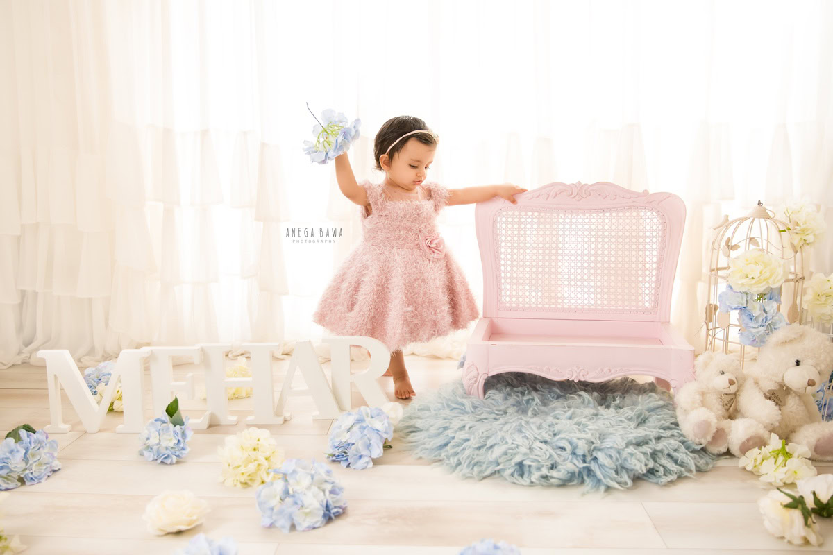 In a captivating photoshoot by Anega Bawa Photography in Delhi and Gurgaon, your 16-month to 1-year-old girl stands gracefully, holding a flower, beside a wooden chair. Against a pristine white backdrop, with a cuddly teddy bear nearby, she exudes innocent charm and creates beautiful memories to cherish forever.