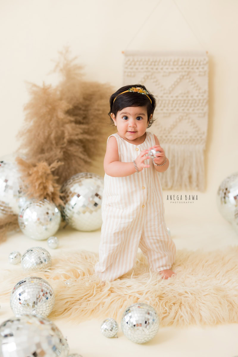 In this photo by Anega Bawa, a 16-month-old girl stands confidently in a jumpsuit, holding a discoball, against a backdrop of brown shrubs and a beige backdrop. With the beige rug under her feet, she exudes a sense of curiosity and exploration, capturing the spirit of toddlerhood in Delhi, Gurgaon, or Noida.