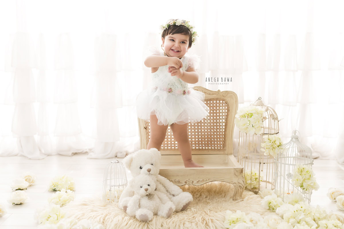Captured by Anega Bawa in Delhi, Gurgaon, this enchanting photo features a 16-month-old girl standing on a wooden chair. Adorned with a tiara band, she stands gracefully on a beige rug amidst white flowers, with a backdrop of serene white. The scene is elevated with the presence of castles, adding a touch of whimsy and charm to the setting.
