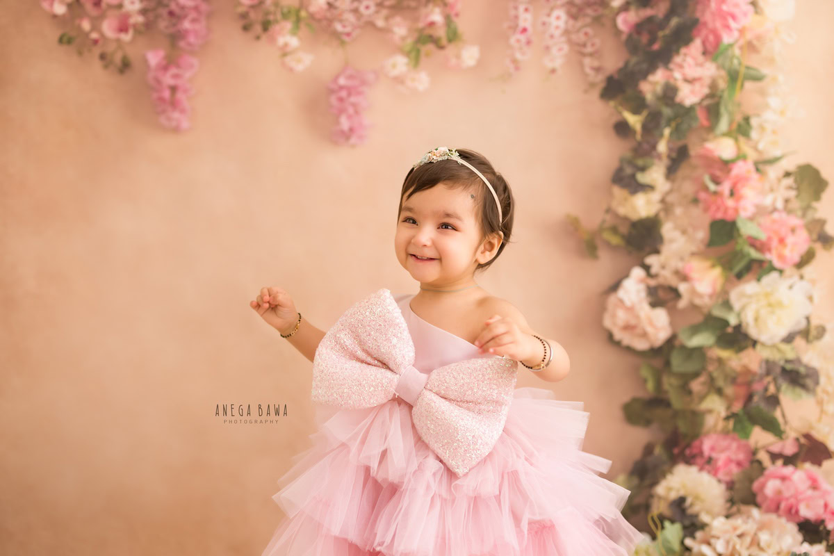 In this delightful photo shoot by Anega Bawa in Delhi, Gurgaon, a 16-month-old girl stands elegantly in a pink bow dress. Against a soft peach backdrop, she radiates sweetness and innocence. The scene is complemented by a floral curve, adding a touch of natural beauty to the setting.