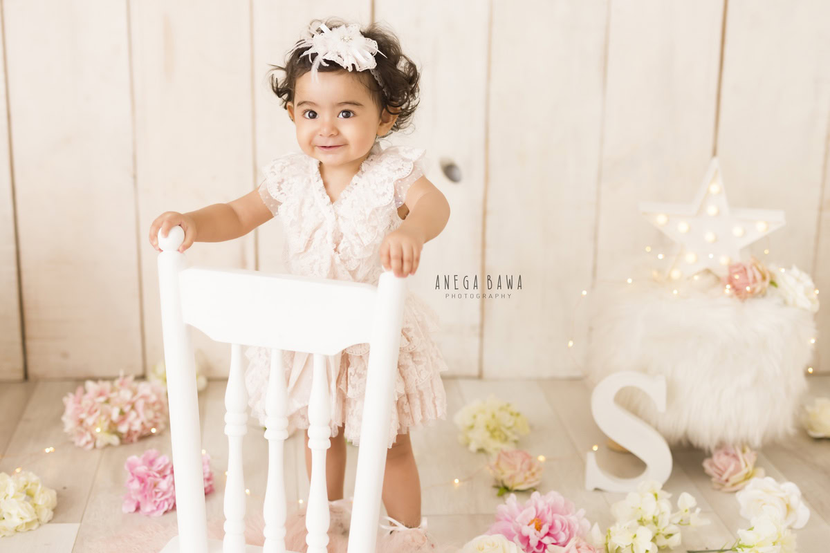 In Gurgaon and Delhi, Anega Bawa captures the innocence of a 17-month-old girl standing beside a white chair against a beige backdrop. Pink and beige flowers add a touch of delicacy to the scene, highlighting her tender age and natural beauty.