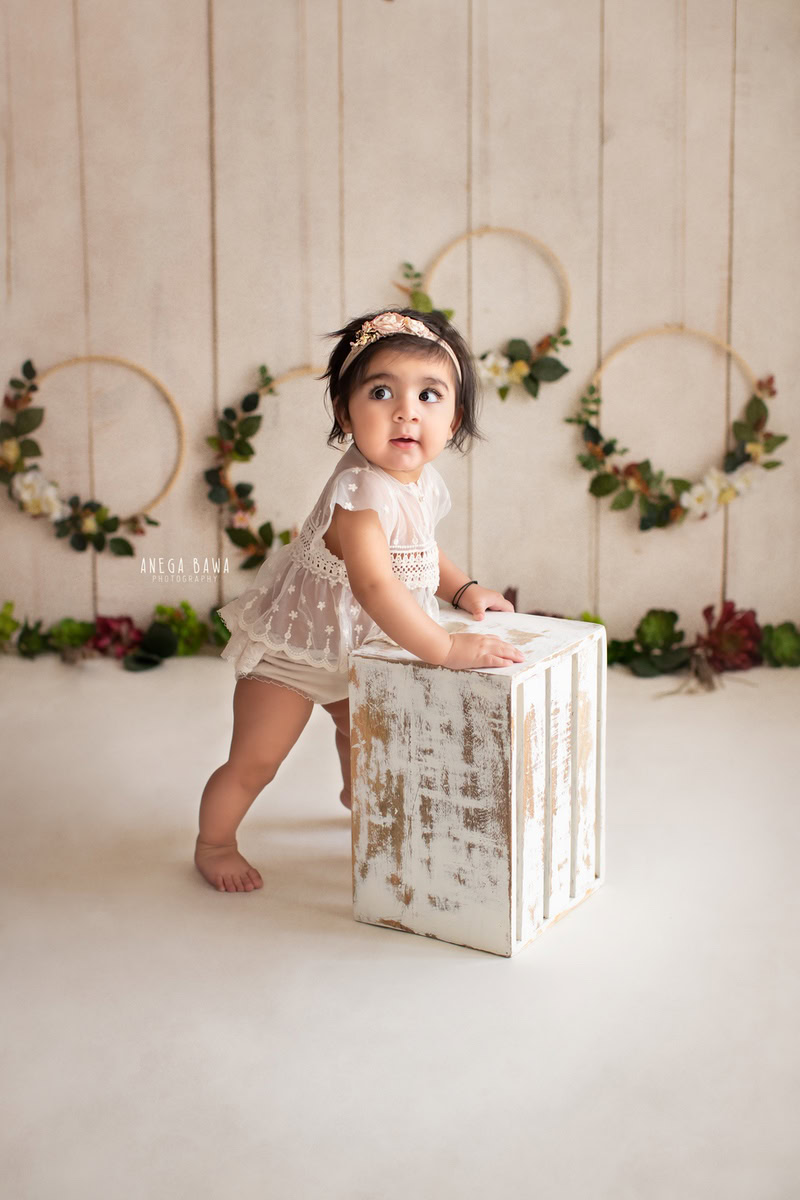 During a baby pre-birthday photoshoot in Delhi, Anega Bawa, Gurgaon, and Noida, a 17-month-old girl stands confidently on a white stool. The beige backdrop serves as a serene canvas, complemented by floral wooden frames adorning the walls. This setup creates a charming atmosphere, capturing the innocence and beauty of the little girl as she strikes a pose.