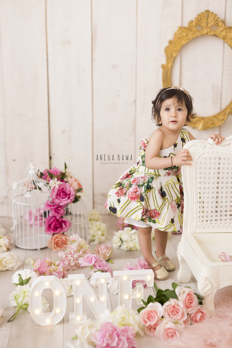 Girl standing beside a white chair against a serene beige backdrop with a golden frame on the wall, surrounded by castles and pink flowers, marking her journey from 17 months to 1 year old during her first birthday photoshoot in Delhi by Anega Bawa, Gurgaon, Noida.