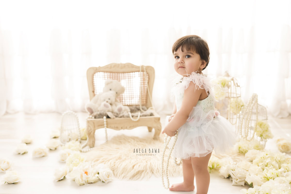 In Gurgaon and Delhi, Anega Bawa frames the enchantment of a 17-month-old girl, standing tall beside a wooden chair against a pristine white backdrop. White flowers and castles add a whimsical touch to the scene, capturing her innocence and grace.