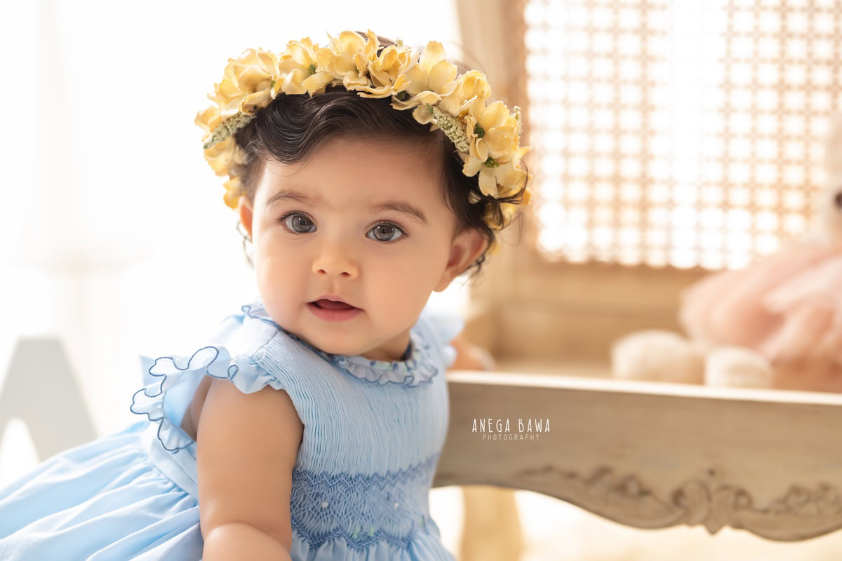 Girl adorned with a tiara band seated on a wooden chair with a teddy bear against a pristine white backdrop, commemorating her journey from 17 months to 1 year old during her first birthday photoshoot in Delhi by Anega Bawa, Gurgaon, Noida.