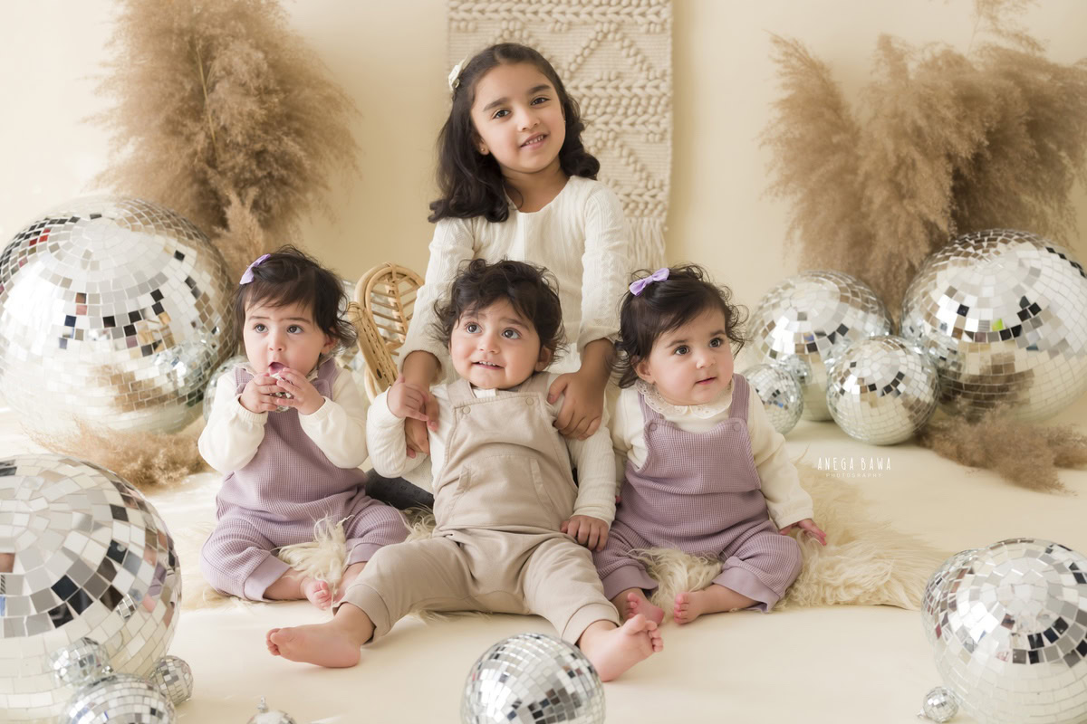 In this charming capture by Anega Bawa, twin siblings, aged 17 months, sit against a backdrop of beige adorned with whimsical discoballs and bushes. This delightful moment celebrates their bond and innocence, perfect for sitter photography in Delhi and Gurgaon.