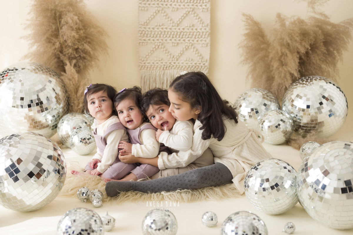Captured by Anega Bawa, this endearing image features twin siblings, aged 17 months, against a backdrop of beige embellished with playful discoballs and bushes. This delightful moment, perfect for sitter photography in Delhi and Gurgaon, captures the joy and bond between the siblings.