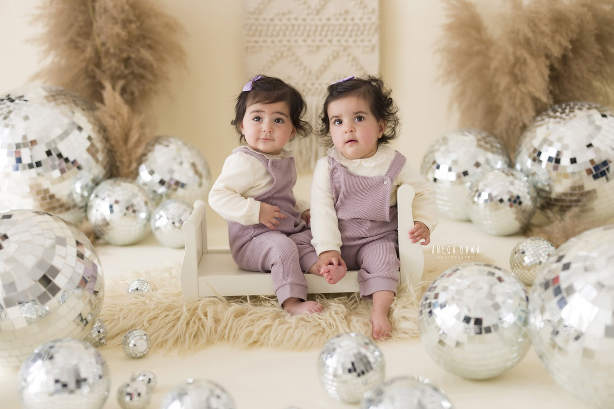In this image captured by Anega Bawa, twin girls, aged 17 months or 1 year old, sit together on a white cot. Behind them, discoballs add a playful touch to the scene, while a beige rug complements the beige backdrop and bushy foliage. The photo captures a moment of shared joy and companionship during a toddler photoshoot in Delhi, Gurgaon, or Noida.