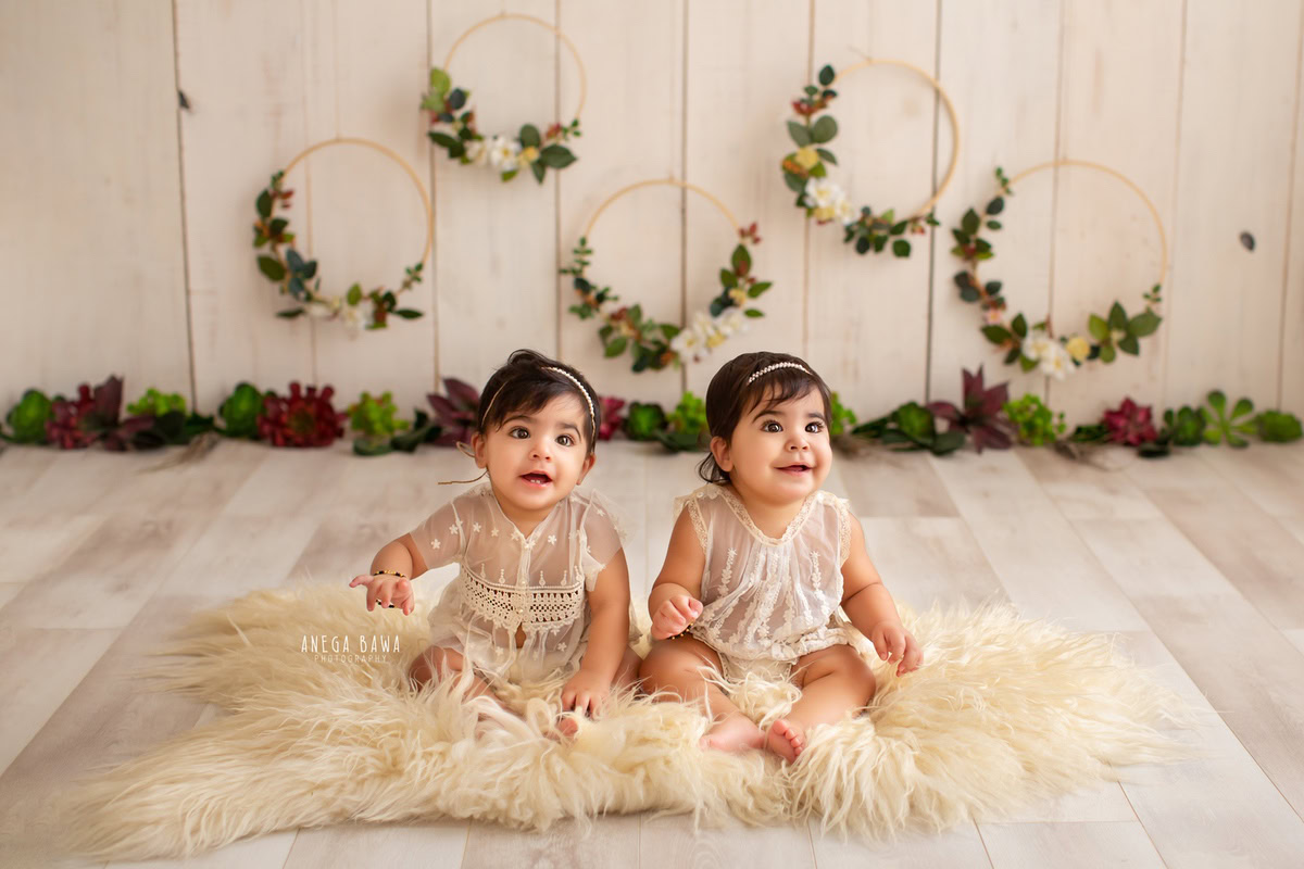 Anega Bawa Photography presents a charming scene in Delhi and Gurgaon, where your 17-month to 1-year-old twin girls, dressed in elegant net dresses, sit on a beige rug. With a floral frame adorning the wall and a soothing beige backdrop, they radiate innocence and grace, capturing a moment of sisterly love in timeless beauty.