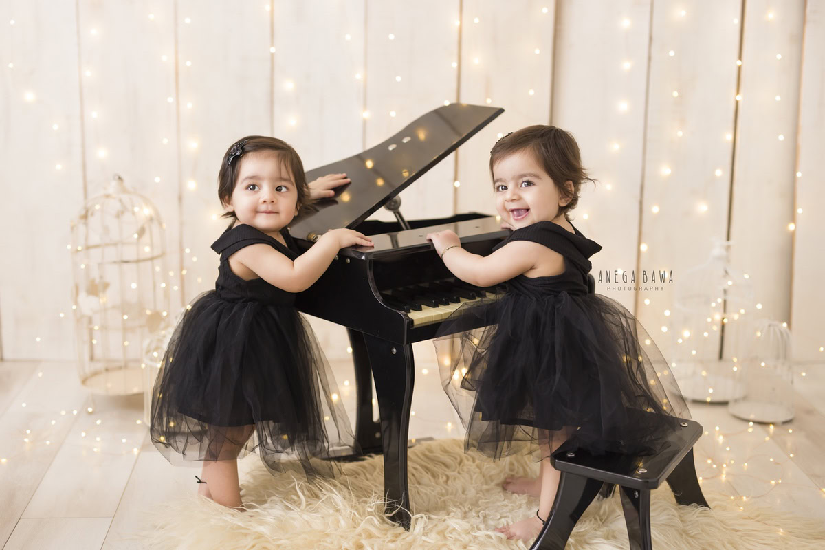 Twin girls playing piano amidst castles against a cozy beige backdrop, illuminated by fairy lights, capturing their joyous journey from 17 months to 1 year old during their first birthday photoshoot in Delhi by Anega Bawa, Gurgaon, Noida.