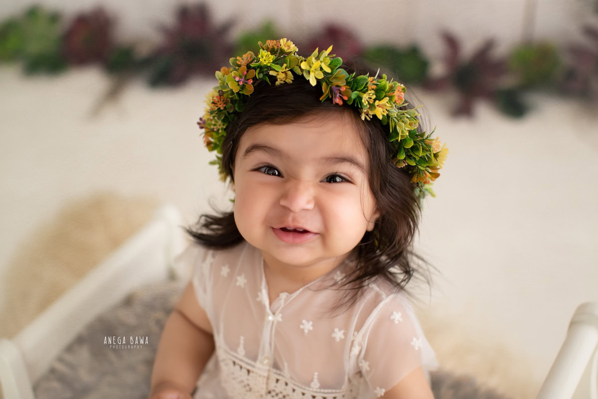 In a delightful photoshoot by Anega Bawa Photography in Delhi and Gurgaon, your 17-month to 1-year-old girl shines in a net dress adorned with a tiara band. Seated gracefully on a grey rug with a charming leafy fringe, she exudes elegance against the backdrop of this enchanting setting.