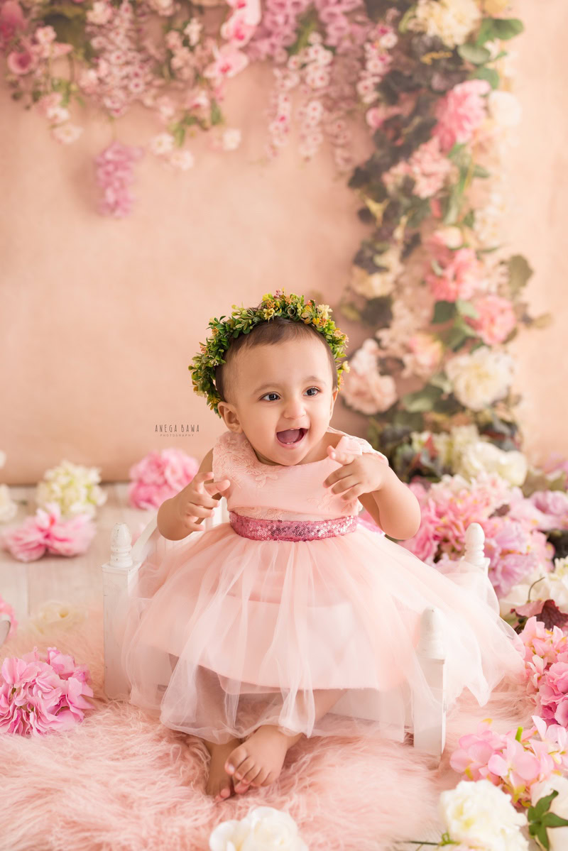 In this adorable toddler photoshoot by Anega Bawa, a 17-month-old girl wears a charming pink dress and a tiara band. She sits on a pink rug surrounded by floral fringes and pink and white flowers, against a peach backdrop. This delightful scene captures the innocence and sweetness of childhood, making it a memorable moment in Delhi, Gurgaon, or Noida.
