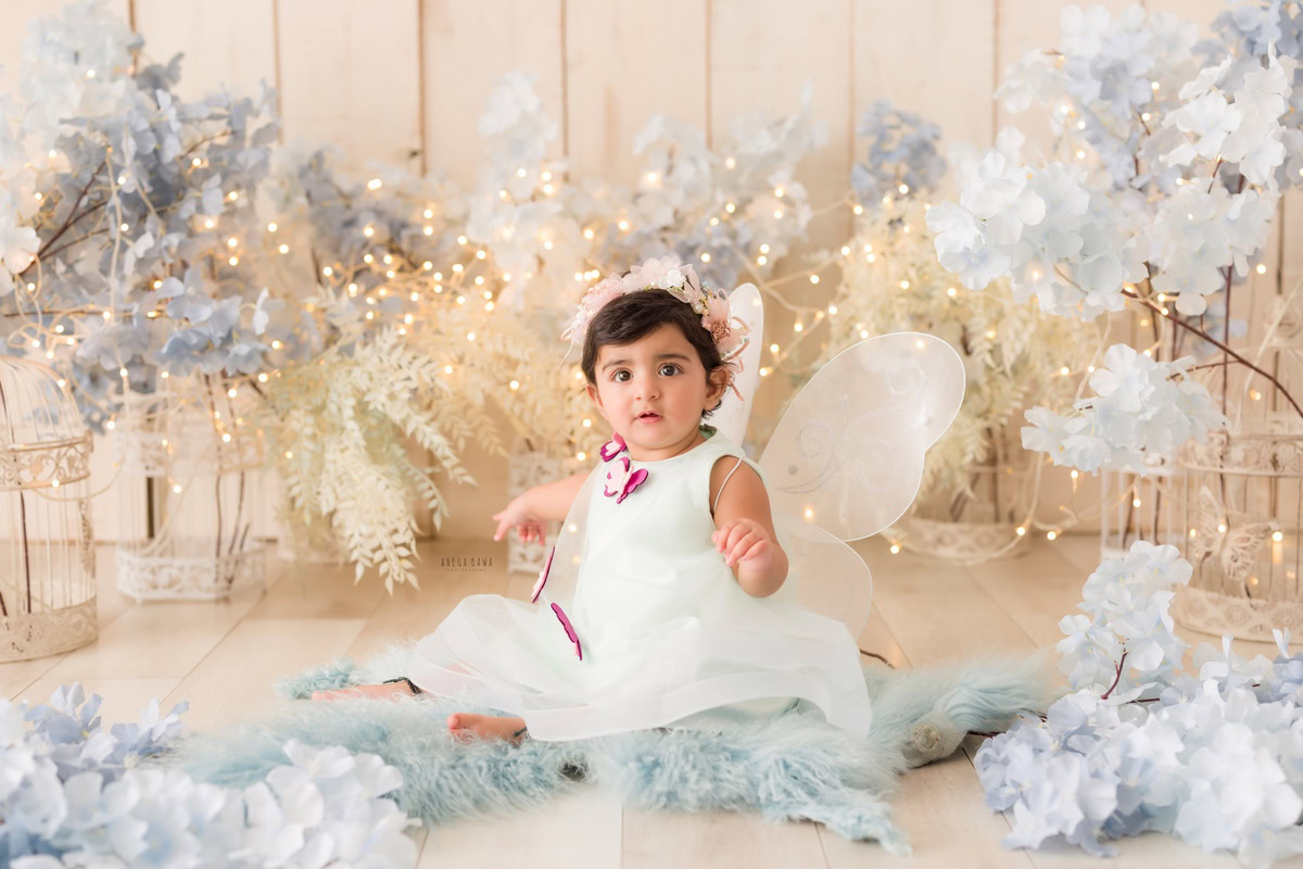 During a baby pre-birthday photoshoot in Delhi, Anega Bawa, Gurgaon, and Noida, a 17-month-old girl dons butterfly wings amidst a backdrop of delicate blue flowers. The beige backdrop, adorned with fairy lights, adds a magical touch to the scene, capturing the enchanting innocence of the little girl in this whimsical setting.