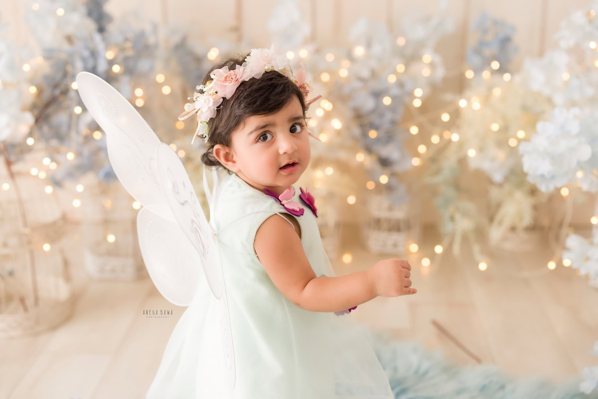 During a baby pre-birthday photoshoot in Delhi, Anega Bawa, Gurgaon, and Noida, a 17-month-old girl poses with butterfly wings on a blue rug surrounded by white and blue flowers. The scene is illuminated with fairy lights, creating a magical ambiance that perfectly captures the innocence and charm of the little girl in this enchanting setting.