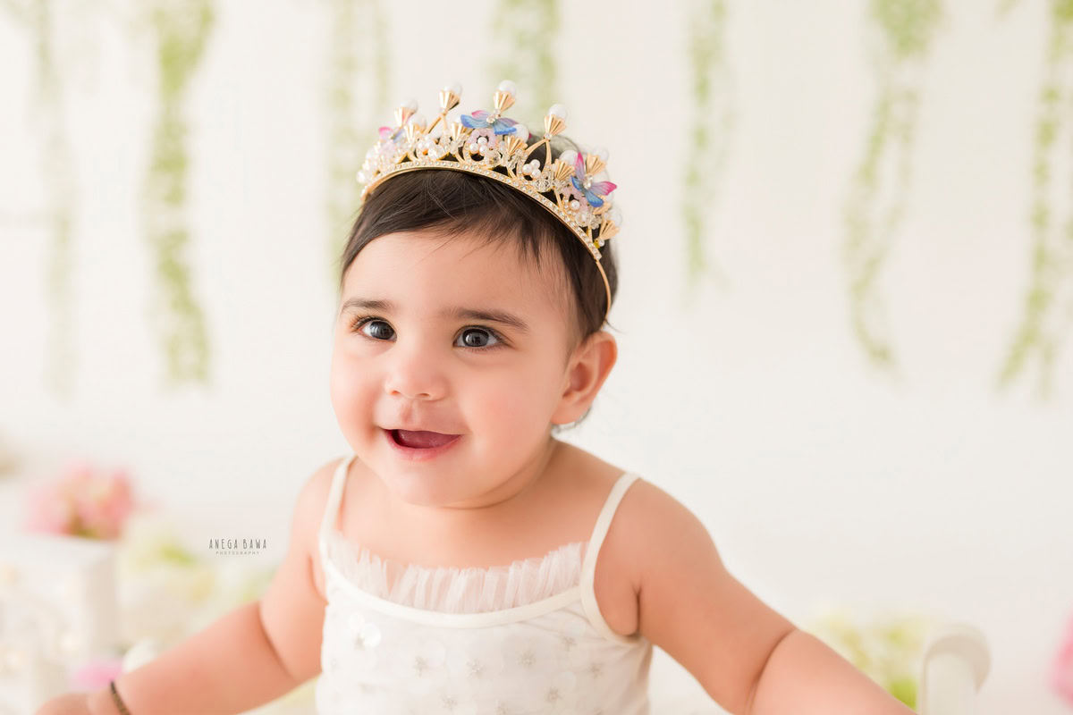 For a baby pre-birthday photoshoot in Delhi, Anega Bawa, Gurgaon, and Noida, a 17-month-old girl wears a crown headband against a leafy fringe and a white backdrop. Her adorable expression, combined with the elegant crown, creates a charming and captivating scene, capturing the essence of her innocence and joy at this special milestone.