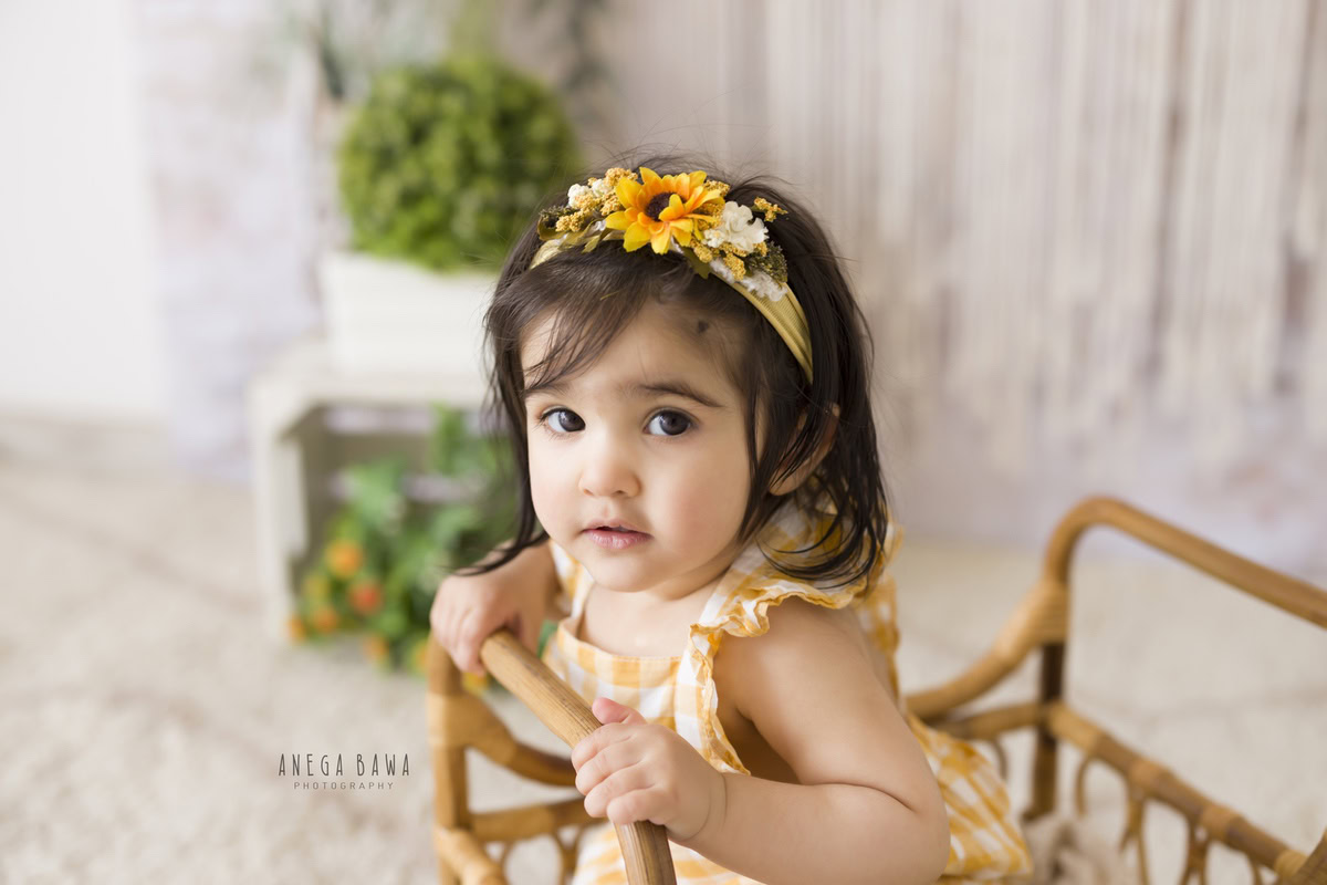 In this charming photo by Anega Bawa, a 17-month-old sits in a wooden cot adorned with a sunflower headband. Against a backdrop of serene white punctuated by white pots, the image captures the innocence and beauty of childhood. Perfect for sitter photography in Delhi and Gurgaon, it encapsulates the tender moments of early life.