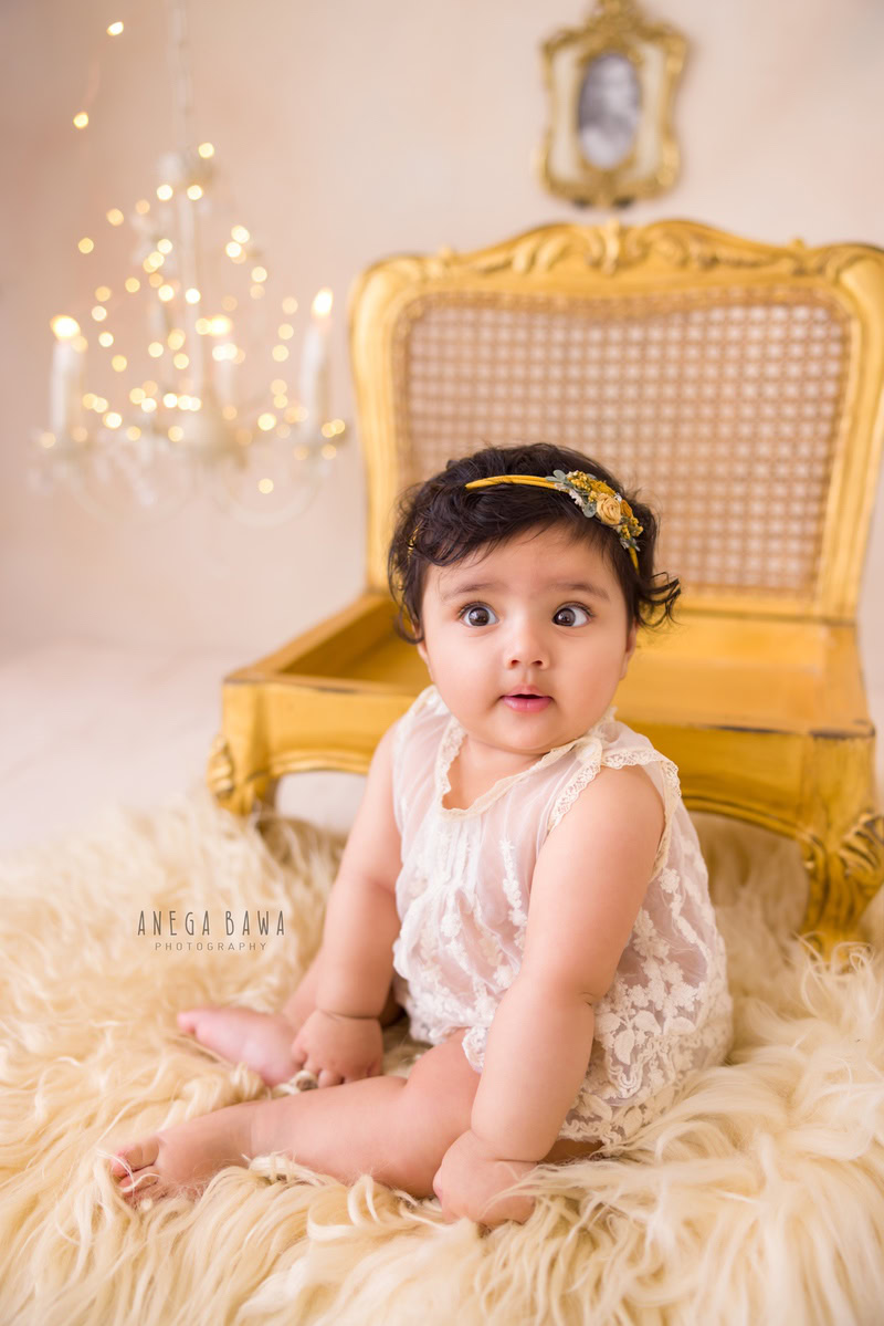 In this enchanting photo session by Anega Bawa in Delhi, Gurgaon, a 17-month-old girl sits gracefully on the floor, adorned with a golden chair. The soft beige rug beneath her feet adds to the warmth of the scene, while golden lights with a candle frame create a magical ambiance.