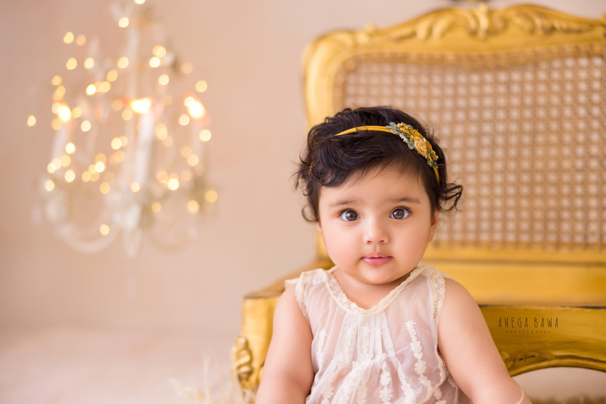 Captured by Anega Bawa in Delhi, Gurgaon, this delightful photo features a 17-month-old girl seated elegantly on the floor beside a golden chair. Adorned with a golden headband, she basks in the glow of golden lights, adding a touch of enchantment to the scene.
