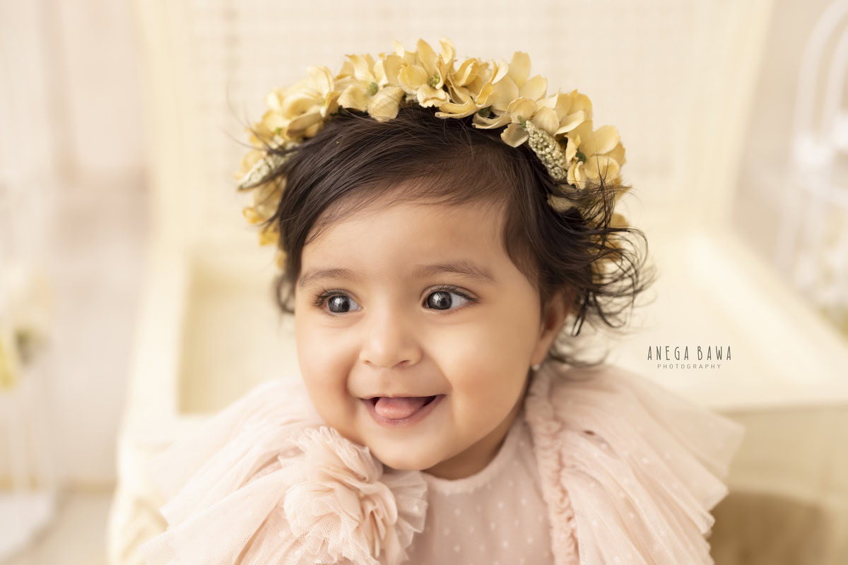 In this enchanting toddler photoshoot by Anega Bawa, a 17-month-old girl adorns a golden tiara band as she sits on a white wooden chair against a pristine white backdrop. This elegant setup beautifully highlights the innocence and charm of the little girl, creating a timeless memory in Delhi, Gurgaon, or Noida.