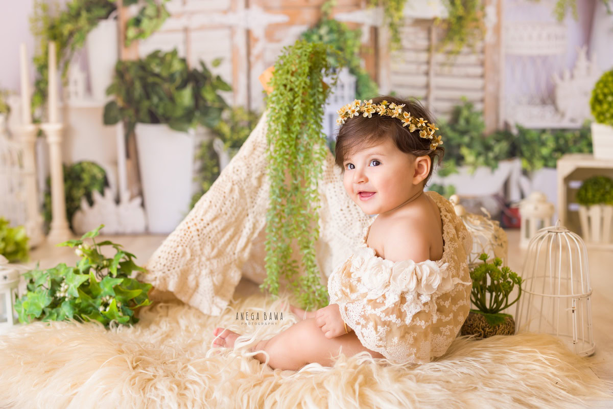 In Gurgaon and Delhi, Anega Bawa captures the serene beauty of a 17-month-old girl seated gracefully on a beige rug, surrounded by white pots adorned with lush green leaves. Dressed in a delicate net dress, she exudes innocence and charm, while white castles add a touch of whimsy to the enchanting scene.