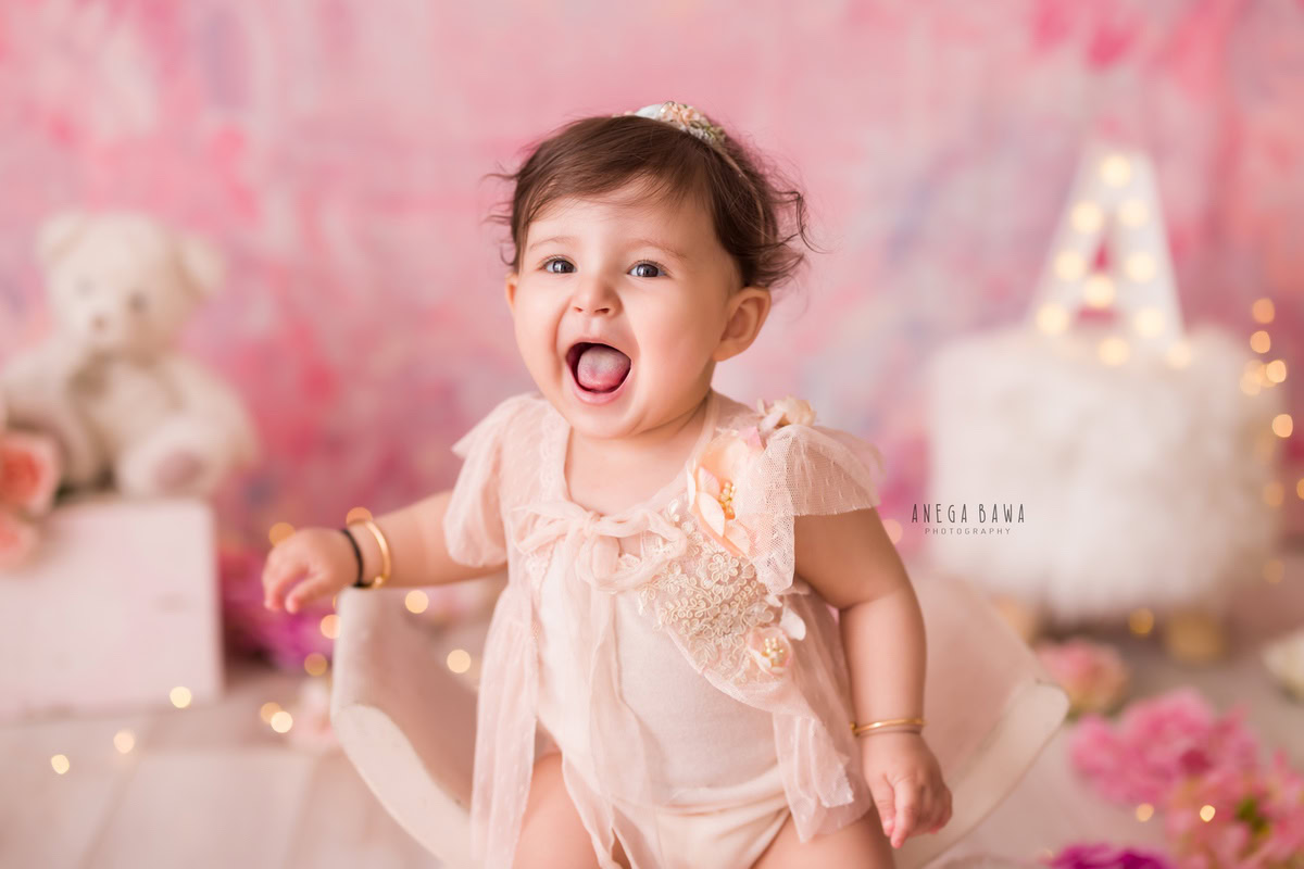 In this charming portrait captured by Anega Bawa in Delhi, Gurgaon, a 1-year-old girl stands against a pink backdrop, exuding innocence and joy. With a teddy bear nestled behind her and surrounded by delicate pink flowers, she adds a touch of sweetness to the scene.
