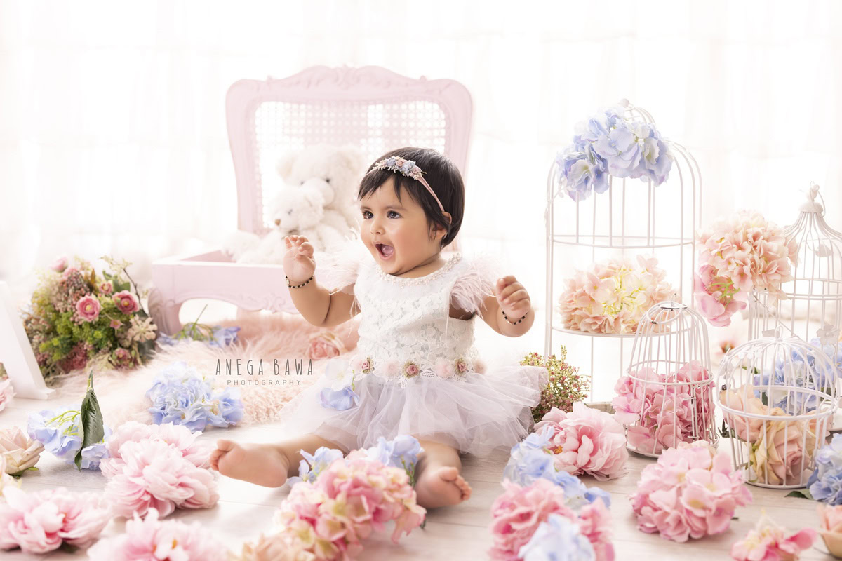 Pink wooden chair adorned with teddy bears, with a girl seated on the floor surrounded by white castles and multicolored flowers against a pristine white backdrop, capturing the charm of her 1-year-old first birthday photoshoot in Delhi by Anega Bawa, Gurgaon, Noida.