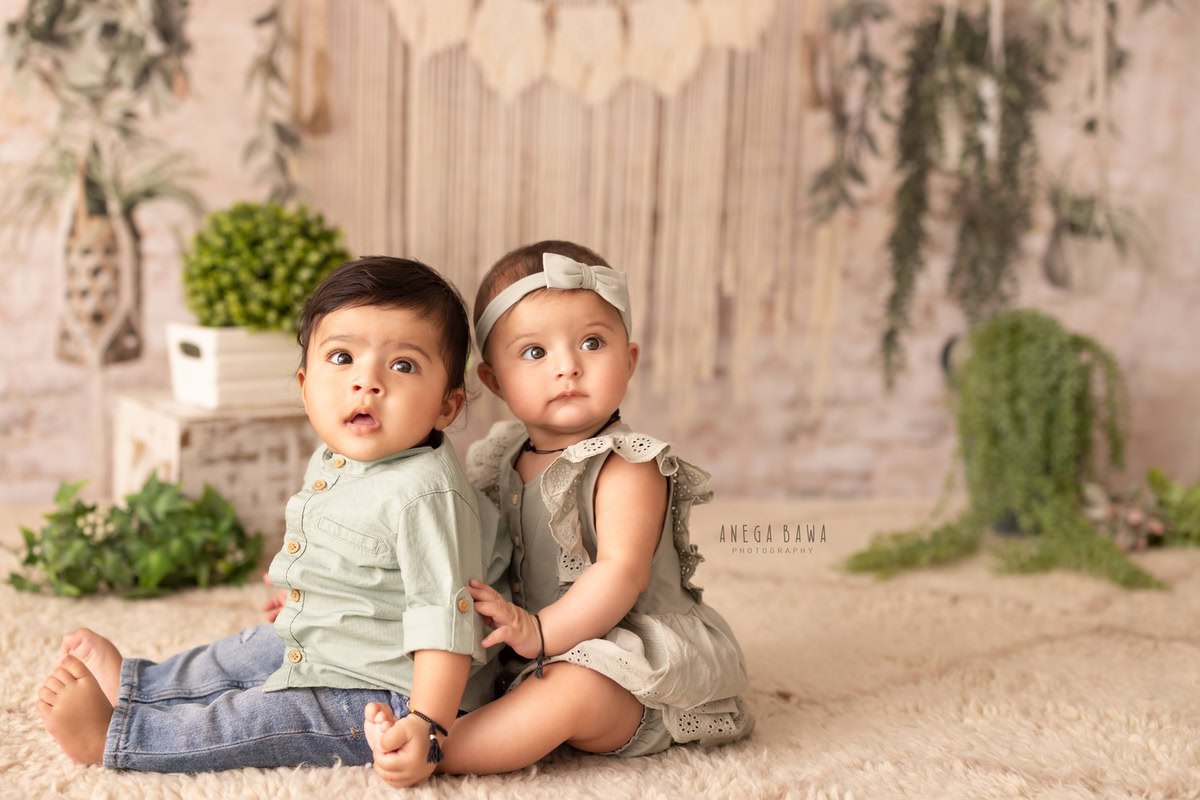 In this endearing shot from Anega Bawa's sitter photoshoot in Delhi, Gurgaon, two siblings are seated on the floor against a soothing beige backdrop. A white vase with leaves falling adds a natural touch to the scene, capturing the innocence and connection between the siblings at 1 year old.
