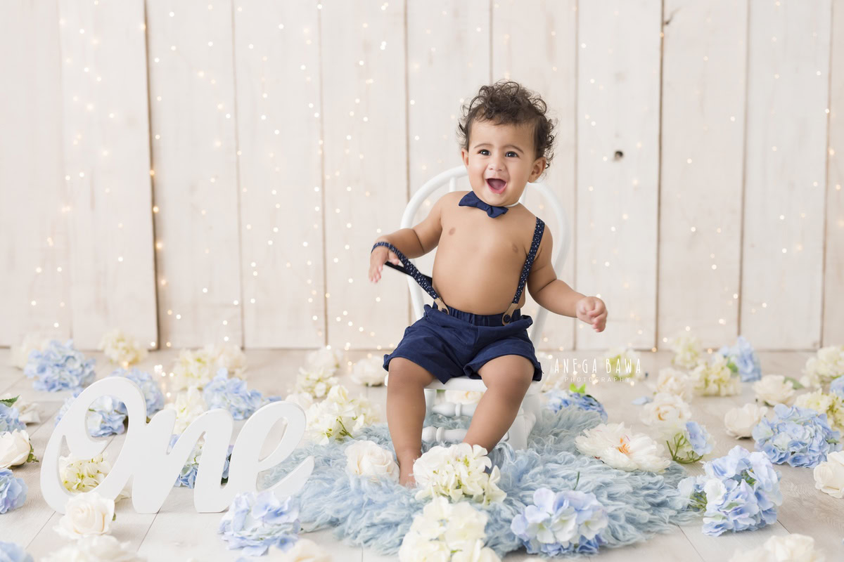 Smiling 1-year-old boy sitting on a chair atop a blue rug surrounded by blue and white flowers, wearing a denim bow and dungaree, radiating joy during his first birthday photoshoot in Delhi by Anega Bawa, Gurgaon, Noida.