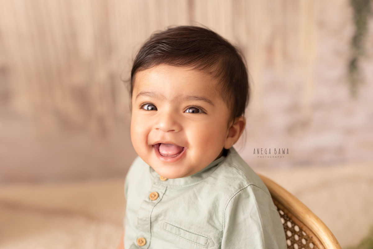 In this delightful capture from Anega Bawa's sitter photoshoot in Delhi, Gurgaon, a smiling boy is seated on a wooden chair against a serene skin backdrop. This candid moment showcases the joy and innocence of the child at 1 year old.