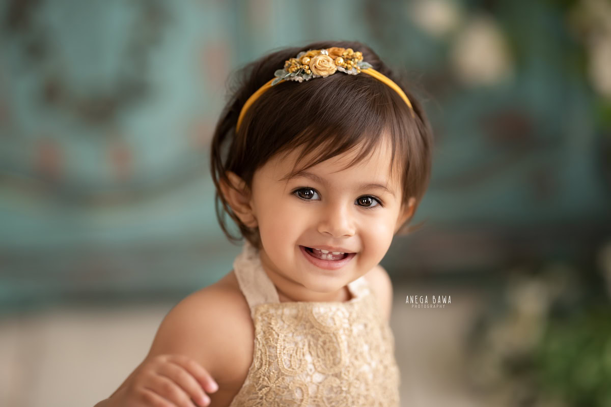 Capture the radiant smile of your 1-year-old girl in a heartwarming photoshoot by Anega Bawa Photography in Delhi and Gurgaon. Adorned with a cute headband, she beams with joy against a charming green backdrop, creating timeless memories to cherish forever.