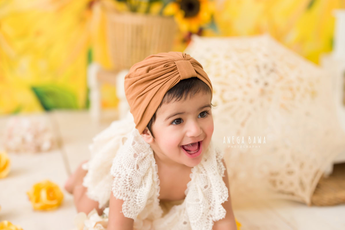 In this adorable toddler photoshoot captured by Anega Bawa in Delhi, Gurgaon, or Noida, a 1-year-old girl poses against a charming sunflower backdrop. She wears a lovely net dress and a brown headband, adding to the sweetness of the scene. A net umbrella on the floor complements the theme, creating a delightful atmosphere for the photoshoot.
