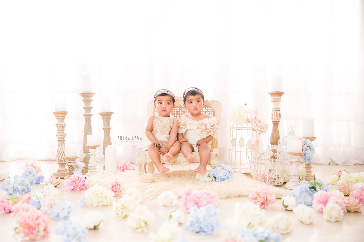 In this charming portrait from Anega Bawa's sitter photoshoot in Delhi, Gurgaon, twin girls are seated on a beige chair next to a candle stand, with flowers adorning the floor. Against a pristine white backdrop, these 1-year-old toddlers radiate innocence and sweetness.