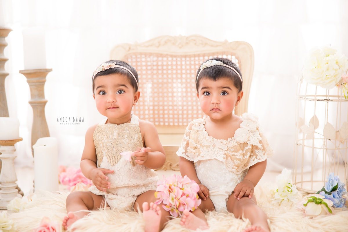 In this delightful image captured during a sitter photo shoot by Anega Bawa in Delhi, Gurgaon, twin girls, both 1 year old, are seated on a beige chair beside a candle stand, surrounded by delicate flowers. Against a clean white backdrop, their innocence and joy shine through.