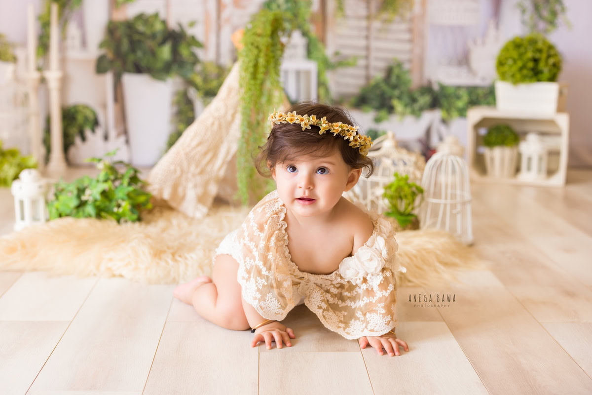 In Gurgaon, Delhi, Anega Bawa captures the innocence and grace of a one-year-old girl in a white backdrop adorned with castles and green leaves. Dressed in a beige net dress and a tiara band, she exudes elegance and charm. White pots with a bunch of leaves add a touch of natural beauty to the scene, creating a whimsical atmosphere captured expertly by the photographer.