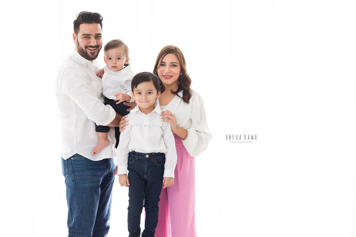 1-year-old boy in dad's arms, posing with his elder brother and mom against a white backdrop, during a family photo shoot by Anega Bawa Photography in Gurgaon (Delhi NCR)