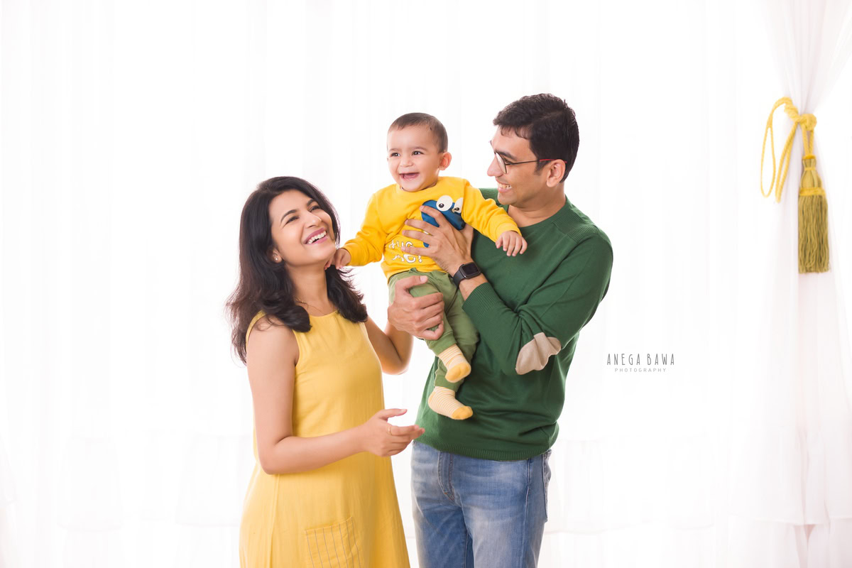 Charming 1-year-old boy in dad's arms, posing with mom against a white backdrop, captured by Anega Bawa Family Photographer Gurgaon (Delhi NCR).