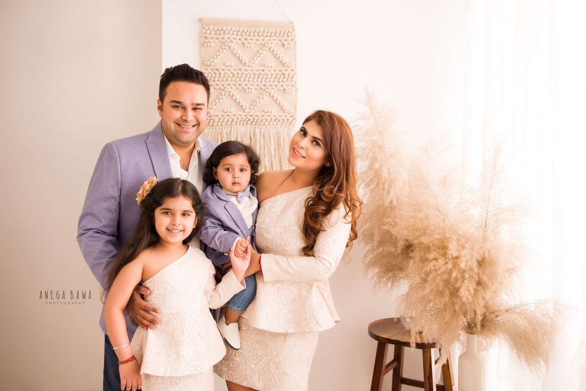 1-year-old boy in mom's arms, posing with elder sister and dad against a backdrop of bushes. This charming family pose was captured by Anega Bawa Photography in Gurgaon (Delhi NCR).