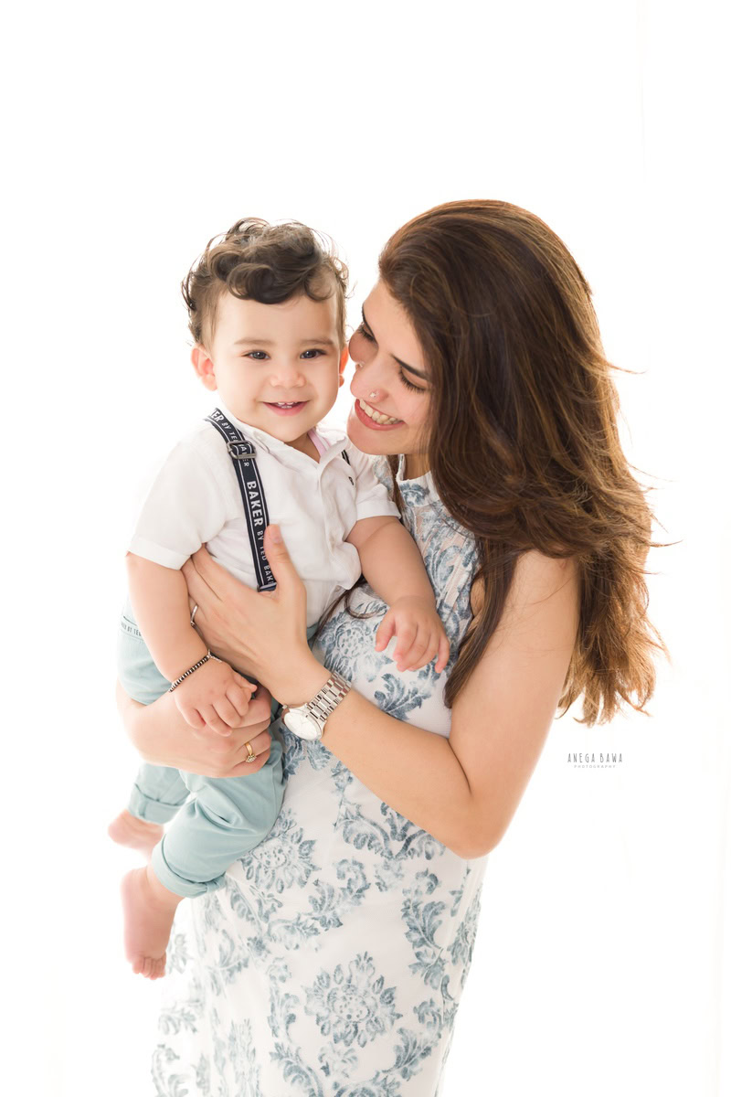 1-year-old boy in mom's arms, posing adorably against a white backdrop. Captured by Anega Bawa Photography, Gurgaon (Delhi NCR).