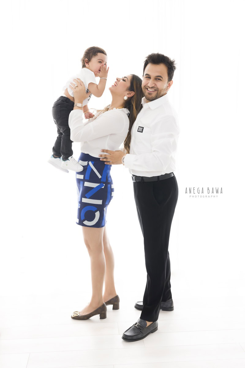 1-year-old boy in mom's arms, posing with dad, all wearing white tees against a white backdrop. This heartwarming family photo was captured by Anega Bawa Photography in Gurgaon (Delhi NCR).