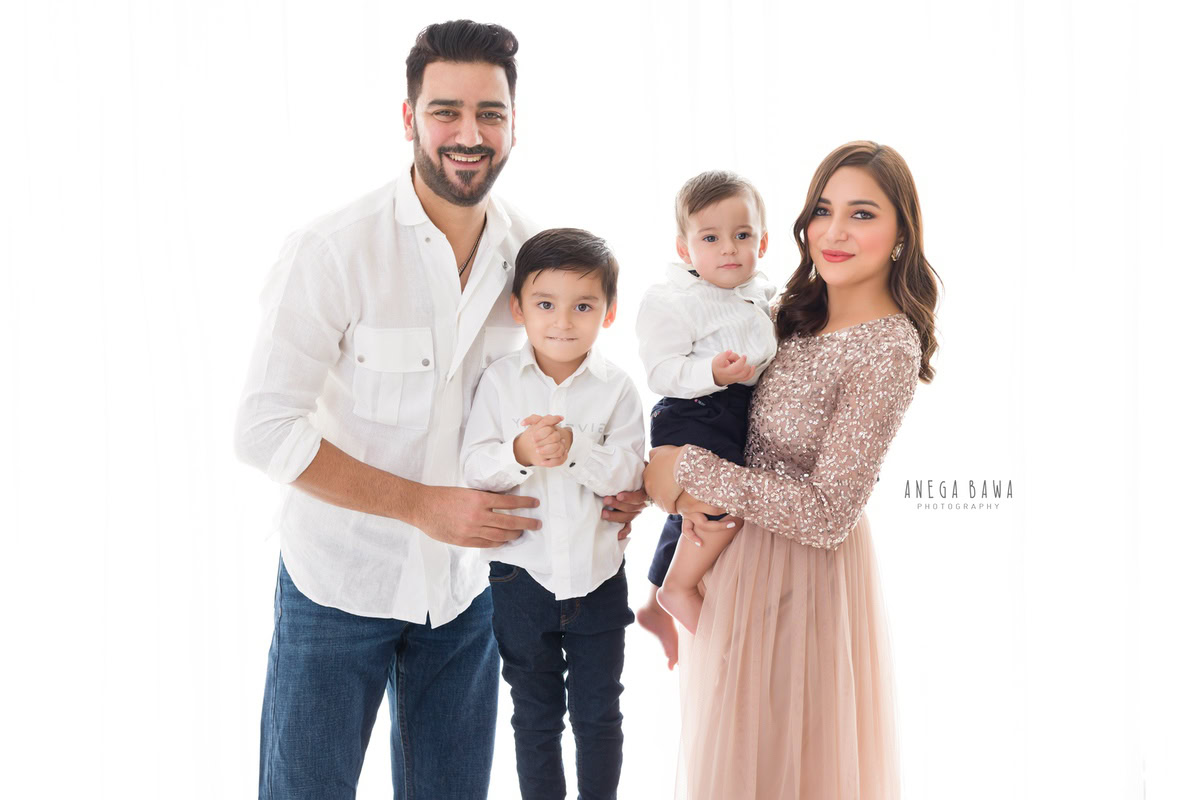 1-year-old boy in mom's arms, posing with dad and elder brother in a family pose against a white backdrop. This beautiful moment was captured by Anega Bawa Photography in Gurgaon (Delhi NCR).