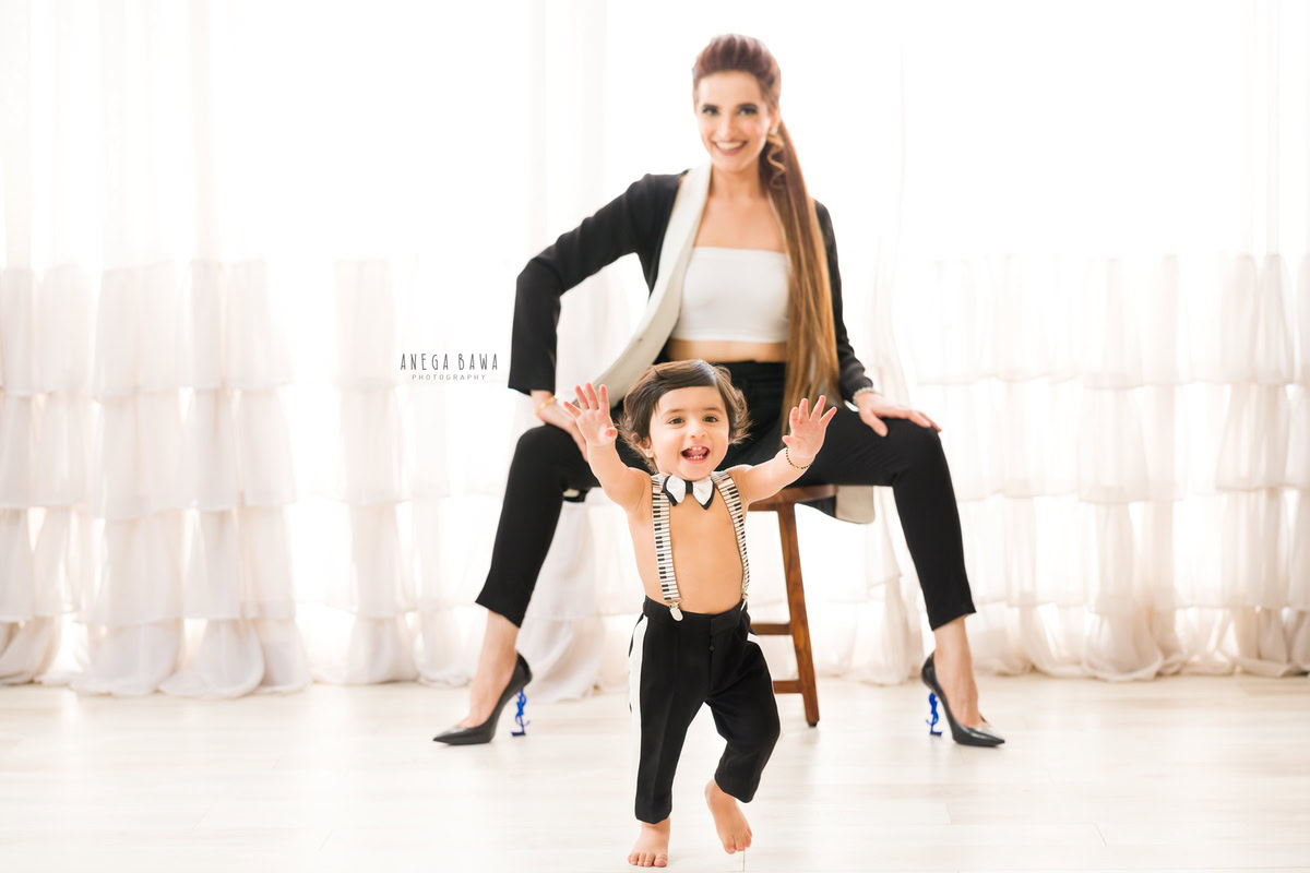 1-year-old boy in a cute bow, posing with his mom seated on a chair against a white backdrop, during a family photo shoot by Anega Bawa Photography in Gurgaon (Delhi NCR)