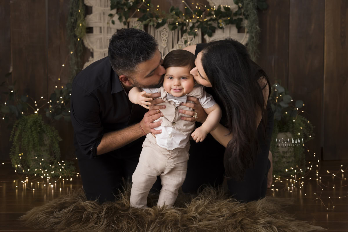 In Gurgaon, Delhi, Anega Bawa beautifully captures a one-year-old boy standing on a brown rug, kissed affectionately by his mom and dad. The scene is enhanced with fairy lights against a warm brown backdrop, creating a tender and memorable moment.