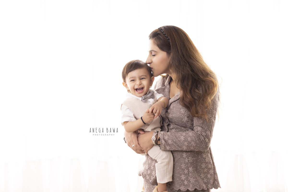 In Gurgaon, Delhi, Anega Bawa captures the heartwarming moment of a one-year-old boy laughing cutely while sharing a kissing pose with his mom against a serene white backdrop.