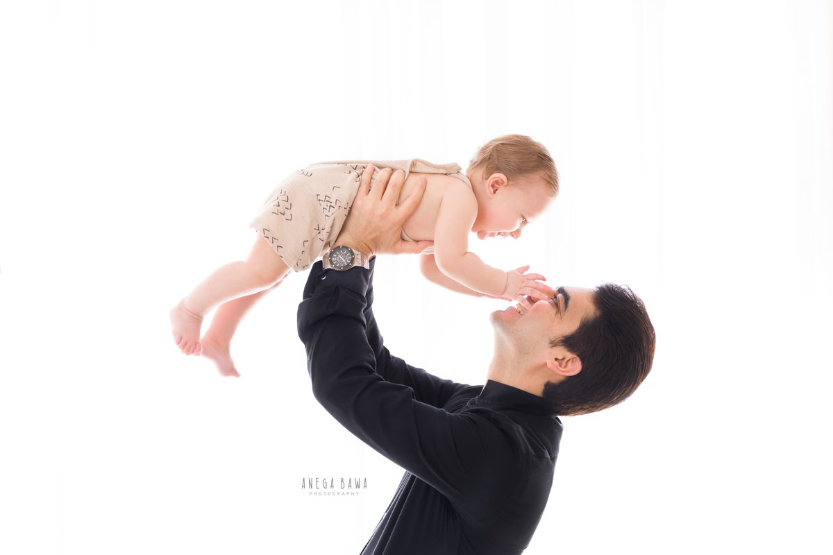 1-year boy lifted in dad's arms against a serene white backdrop, captured by Anega Bawa Photography in Gurgaon (Delhi NCR).